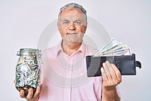 Senior grey-haired man holding wallet with dollars and jar with savings smiling with a happy and cool smile on face