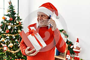 Senior grey-haired man talking on the smartphone holding christmas gift at home