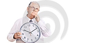Senior grey-haired man wearing business clothes and holding clock serious face thinking about question with hand on chin,