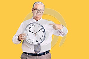 Senior grey-haired man wearing business clothes and holding clock smiling happy pointing with hand and finger