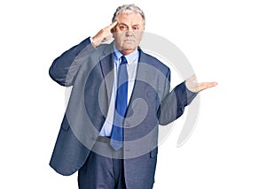 Senior grey-haired man wearing business jacket confused and annoyed with open palm showing copy space and pointing finger to