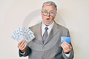 Senior grey-haired man wearing business suit holding credit car and dollars in shock face, looking skeptical and sarcastic,