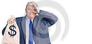 Senior grey-haired man wearing business suit holding money bag with dollar symbol smiling happy doing ok sign with hand on eye