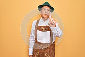 Senior grey-haired man wearing german traditional octoberfest suit over yellow background Pointing with finger up and angry