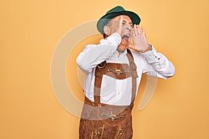 Senior grey-haired man wearing german traditional octoberfest suit over yellow background Shouting angry out loud with hands over