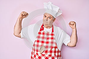 Senior grey-haired man wearing professional baker apron showing arms muscles smiling proud