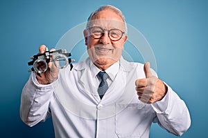 Senior grey haired optic doctor man holding optometrist eyeglasses over blue background happy with big smile doing ok sign, thumb