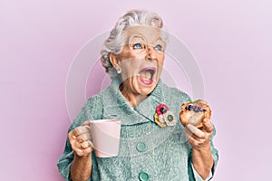 Senior grey-haired woman drinking a cup of coffee and eating bun angry and mad screaming frustrated and furious, shouting with