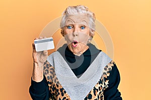 Senior grey-haired woman holding credit card scared and amazed with open mouth for surprise, disbelief face