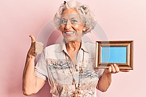 Senior grey-haired woman holding empty frame smiling happy and positive, thumb up doing excellent and approval sign