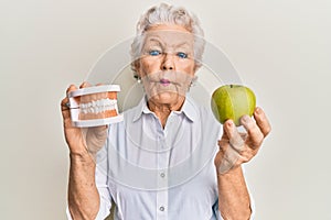 Senior grey-haired woman holding green apple and denture teeth making fish face with mouth and squinting eyes, crazy and comical