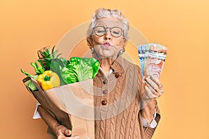 Senior grey-haired woman holding groceries and canadian dollars looking at the camera blowing a kiss being lovely and sexy