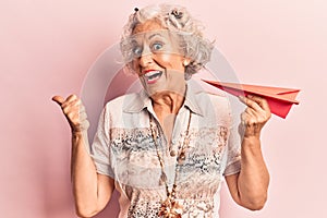 Senior grey-haired woman holding paper airplane pointing thumb up to the side smiling happy with open mouth