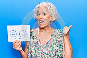 Senior grey-haired woman holding sad to happy emotion paper pointing thumb up to the side smiling happy with open mouth