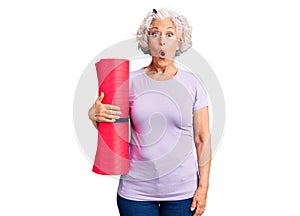 Senior grey-haired woman holding yoga mat scared and amazed with open mouth for surprise, disbelief face