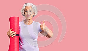 Senior grey-haired woman holding yoga mat smiling happy and positive, thumb up doing excellent and approval sign