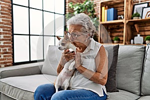 Senior grey-haired woman kissing and hugging chiuahua sitting on sofa at home