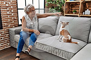 Senior grey-haired woman playing with chiuahua sitting on sofa at home