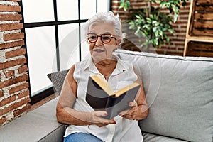 Senior grey-haired woman reading bible sitting on sofa at home