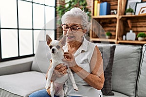 Senior grey-haired woman smiling confident holding chiuahua sitting on sofa at home