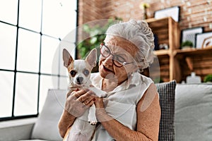 Senior grey-haired woman smiling confident holding chiuahua sitting on sofa at home