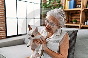 Senior grey-haired woman smiling confident holding chiuahua sitting on sofa at home