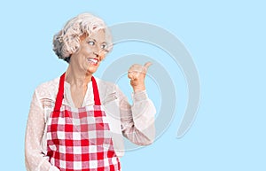 Senior grey-haired woman wearing apron smiling with happy face looking and pointing to the side with thumb up