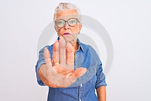 Senior grey-haired woman wearing denim shirt and glasses over isolated white background doing stop sing with palm of the hand