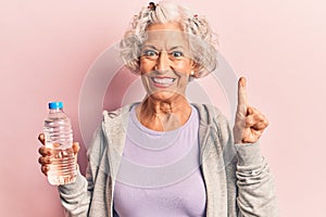 Senior grey-haired woman wearing sportswear drinking bottle of water smiling with an idea or question pointing finger with happy