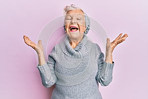 Senior grey-haired woman wearing wool sweater and winter hat celebrating mad and crazy for success with arms raised and closed