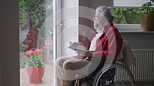 Senior handicapped man in wheelchair reading book looking out the window on rainy day. Portrait of intelligent disabled