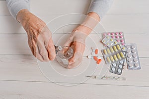 Senior hands with medicine bottle putting drops in a glass, pills, drugs, thermometer on a table.