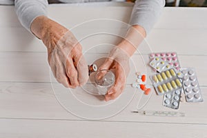 Senior hands with medicine bottle putting drops in a glass, pills, drugs, thermometer on a table.