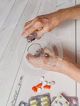 Senior hands with medicine bottle putting drops in a glass, pills, drugs, thermometer on a table.