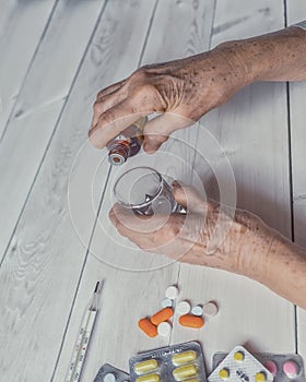 Senior hands with medicine bottle putting drops in a glass, pills, drugs, thermometer on a table.