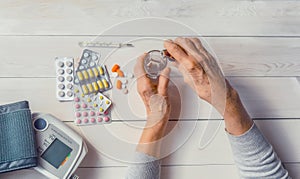 Senior hands with medicine bottle putting drops in a glass, pills, tonometer, thermometer on a table.