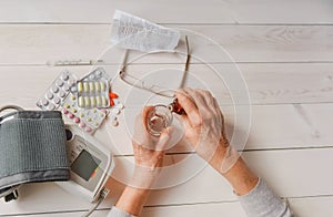 Senior hands with medicine bottle putting drops in a glass, pills, tonometer, thermometer on a table.