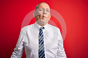 Senior handsome businessman wearing elegant tie standing over isolated red background making fish face with lips, crazy and