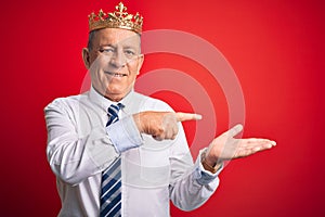 Senior handsome businessman wearing king crown standing over isolated red background amazed and smiling to the camera while