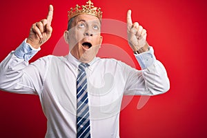 Senior handsome businessman wearing king crown standing over isolated red background amazed and surprised looking up and pointing