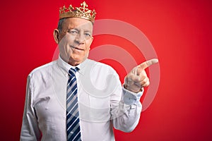 Senior handsome businessman wearing king crown standing over isolated red background with a big smile on face, pointing with hand