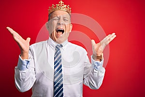 Senior handsome businessman wearing king crown standing over isolated red background celebrating mad and crazy for success with