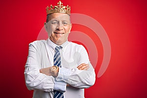 Senior handsome businessman wearing king crown standing over isolated red background happy face smiling with crossed arms looking