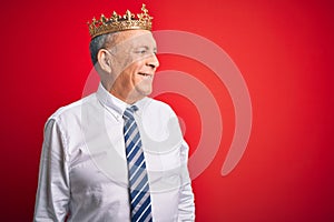 Senior handsome businessman wearing king crown standing over isolated red background looking away to side with smile on face,