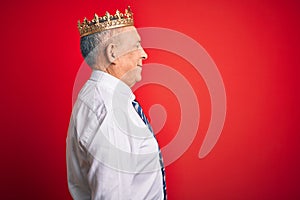 Senior handsome businessman wearing king crown standing over isolated red background looking to side, relax profile pose with