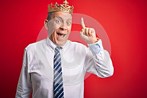 Senior handsome businessman wearing king crown standing over isolated red background pointing finger up with successful idea