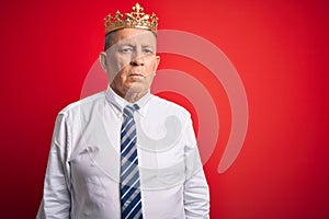 Senior handsome businessman wearing king crown standing over isolated red background with serious expression on face