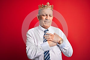 Senior handsome businessman wearing king crown standing over isolated red background smiling with hands on chest with closed eyes