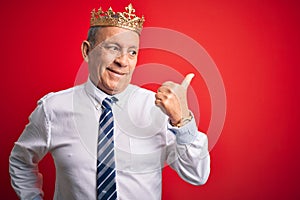 Senior handsome businessman wearing king crown standing over isolated red background smiling with happy face looking and pointing