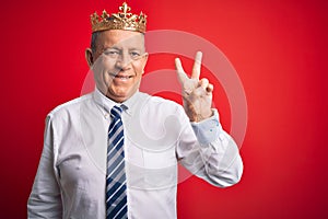 Senior handsome businessman wearing king crown standing over isolated red background smiling with happy face winking at the camera